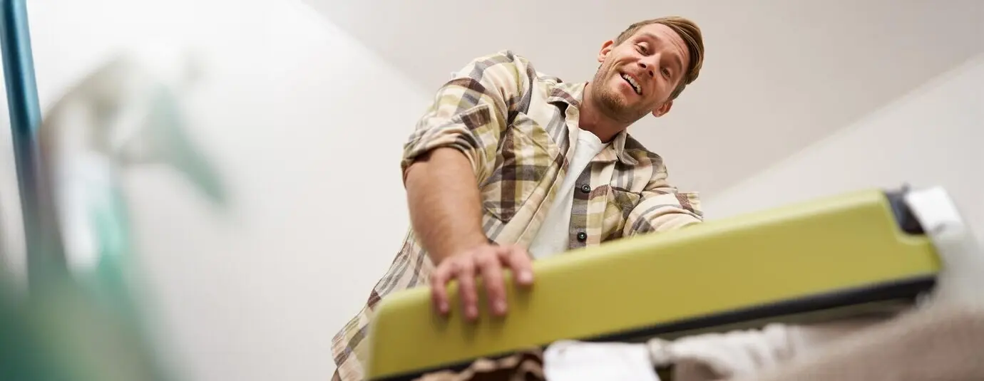 Porträt aus der Untersicht eines jungen, muskulösen Mannes, der versucht, seinen mit Kleidung gefüllten Koffer zu schließen, indem er mit Kraft darauf drückt.